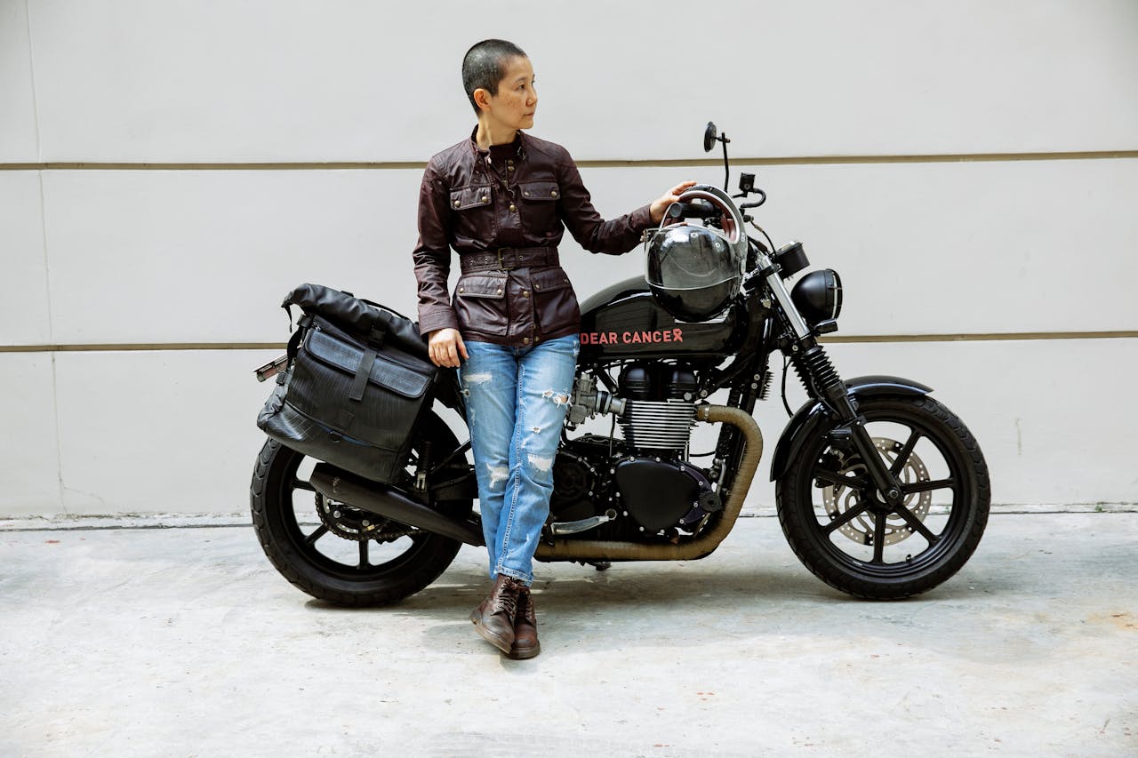 Woman in riding gear standing beside a black motorcycle labeled "Dear Cancer," holding a helmet and leaning confidently against the tank.