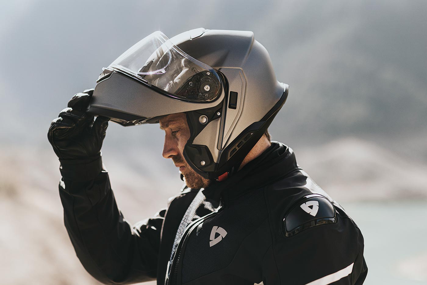 Rider lifting the chin bar of a matte modular helmet in a mountain setting