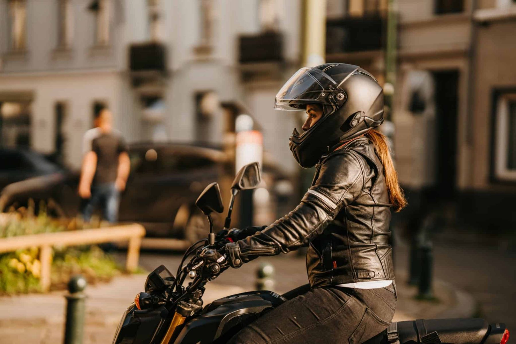 Woman in a black helmet and leather jacket riding a motorcycle in the city, gripping the handlebars with proper form.