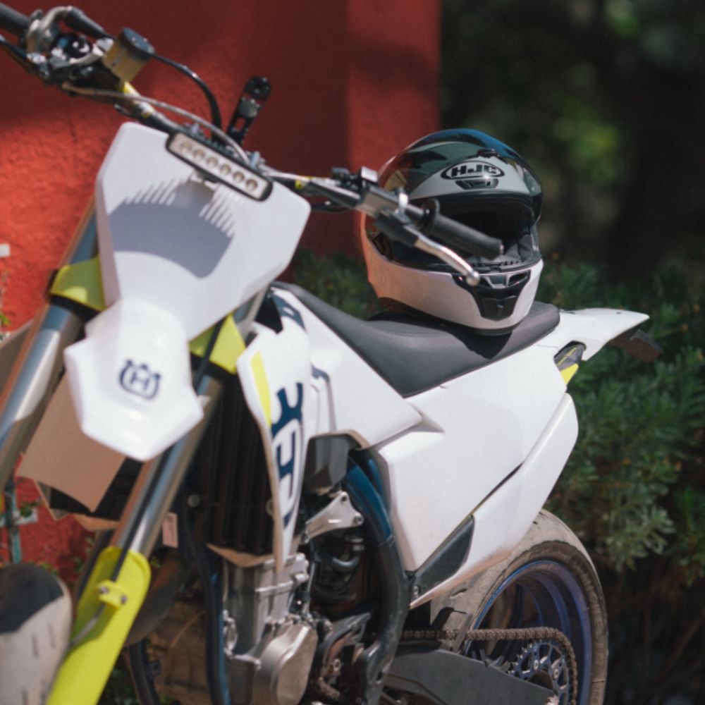 HJC i10 full-face helmet resting on the seat of a Husqvarna motorcycle, showing its compact design and Snell-certified protection.