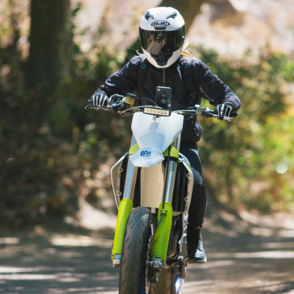 Rider wearing an HJC i10 full-face helmet with tinted visor, riding a Husqvarna motorcycle on a shaded forest road.