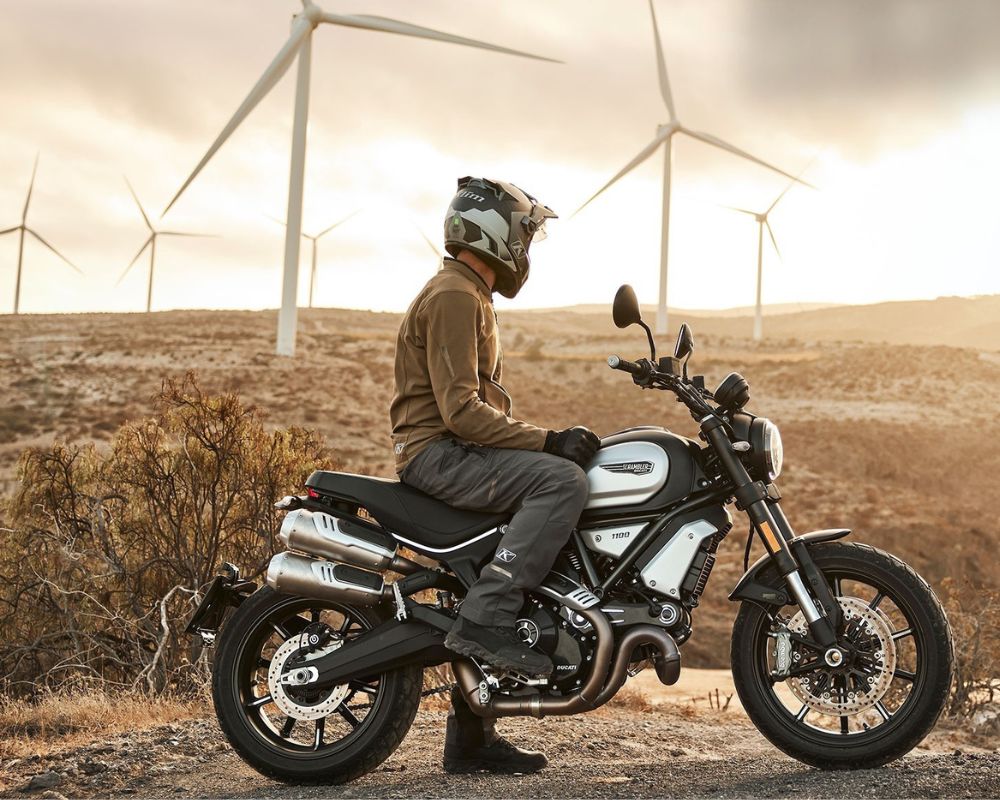 Rider wearing Klim Marrakesh jacket on Ducati Scrambler in desert landscape with wind turbines.