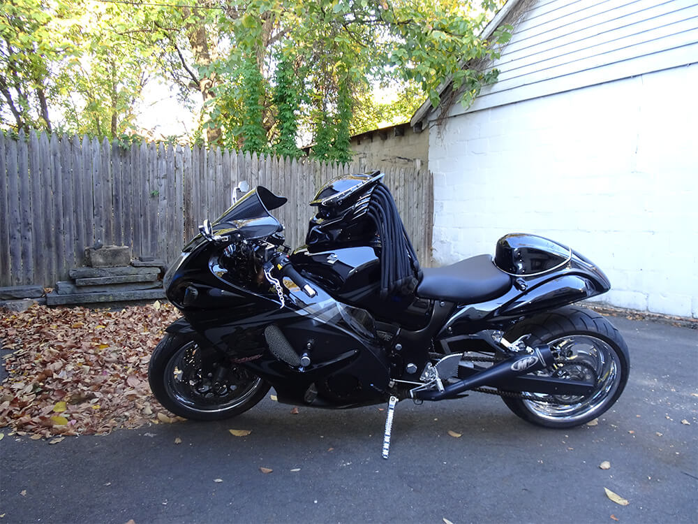 Custom motorcycle with a Predator-style helmet mounted on the tank, featuring dreadlocks and blacked-out design.