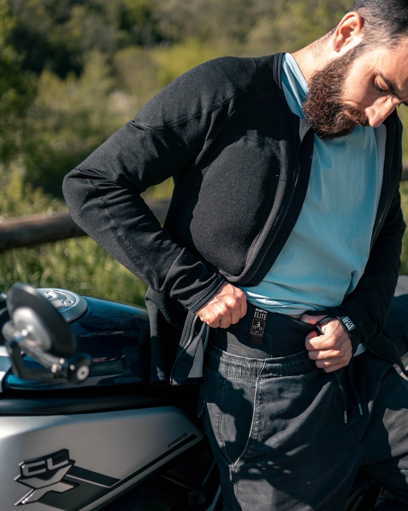 Rider adjusting Bowtex Elite leggings waistband next to a motorcycle, showing minimalist design with deep internal armour pockets.