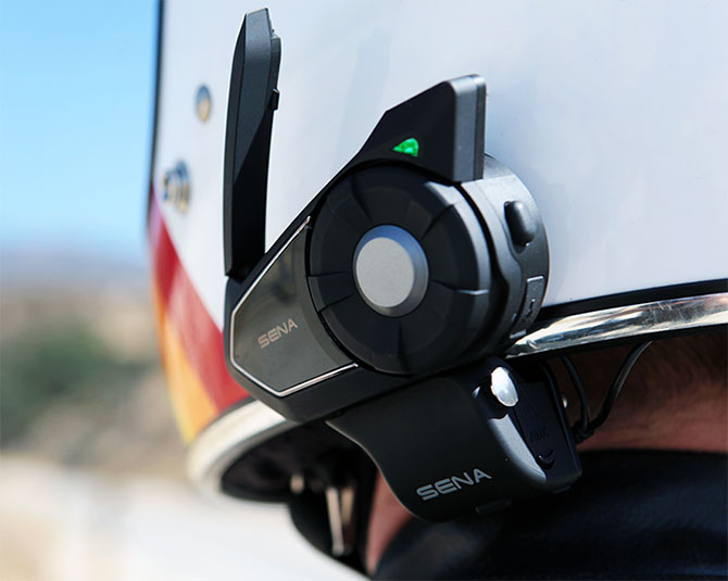 Close-up of Sena 30K Bluetooth headset mounted on a motorcycle helmet, antenna raised and green status light visible.