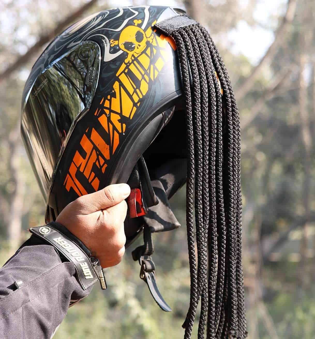 Motorcyclist holding a custom helmet with attached Predator-style dreadlocks and orange skull graphics, outdoors in sunlight.