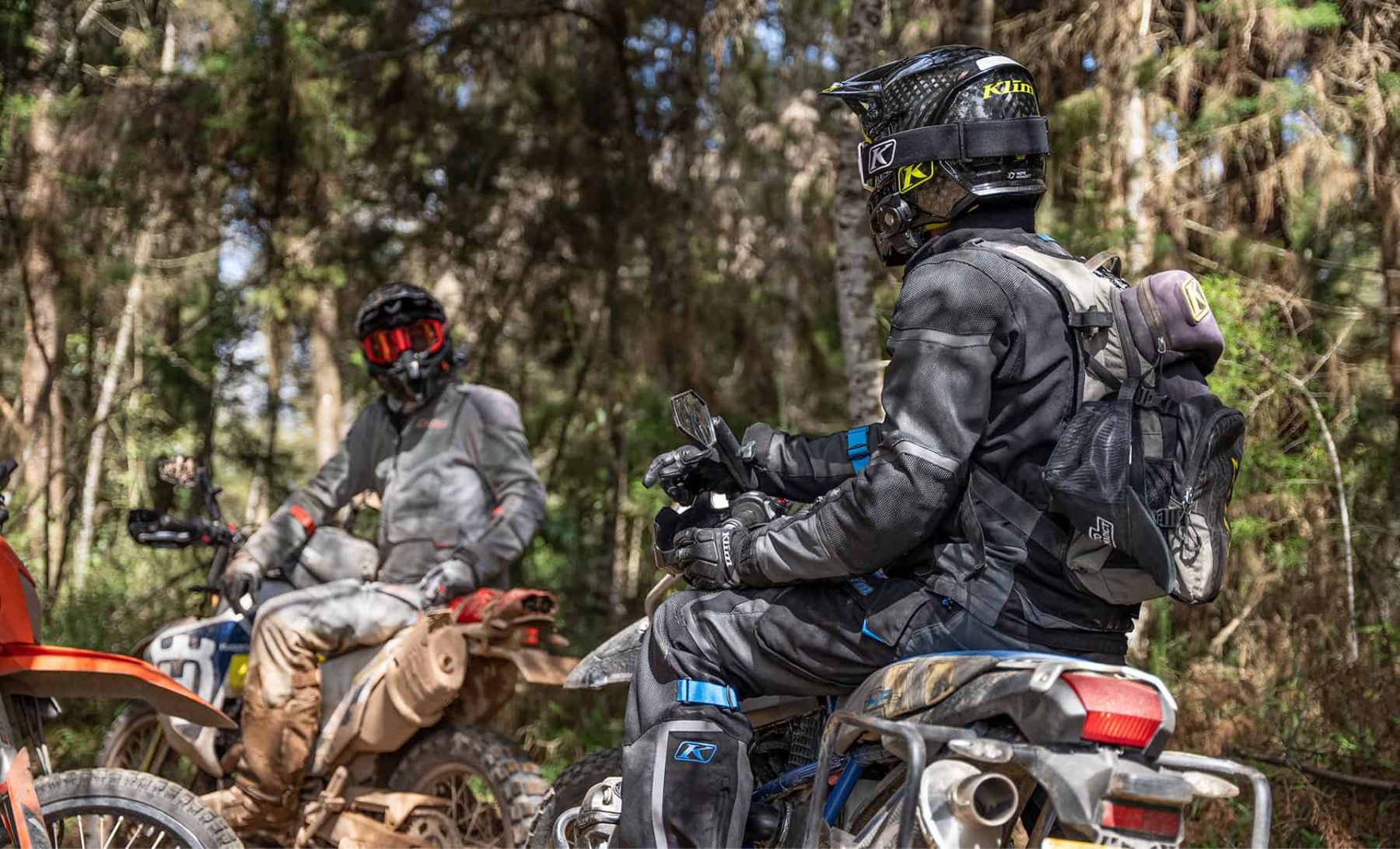 Two ADV riders wearing Klim Baja S4 jackets stopped in a forest trail, conversing on their motorcycles