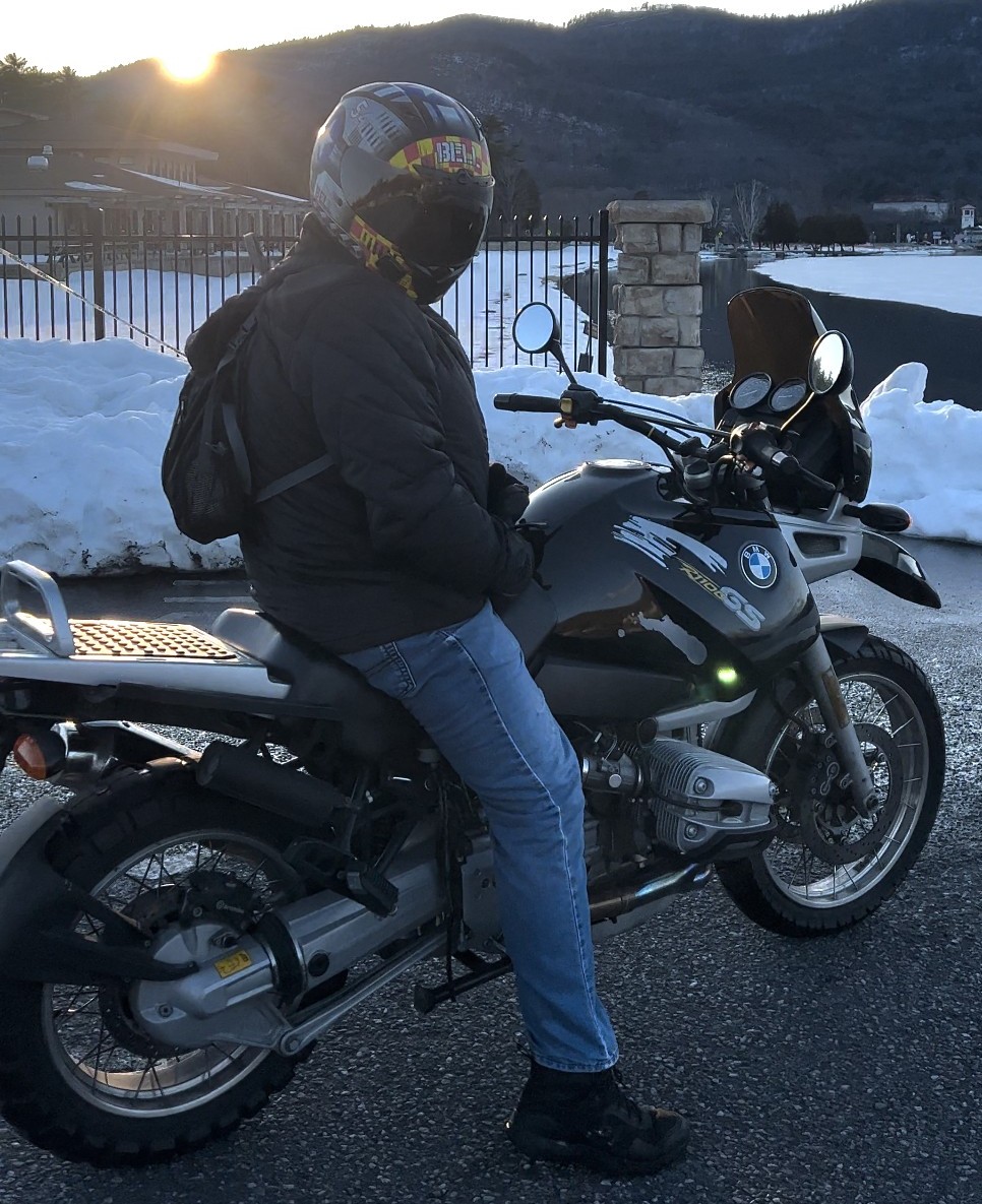 Rider wearing a Bell Qualifier DLX MIPS helmet on a BMW GS motorcycle, parked by snowy mountains at sunset.