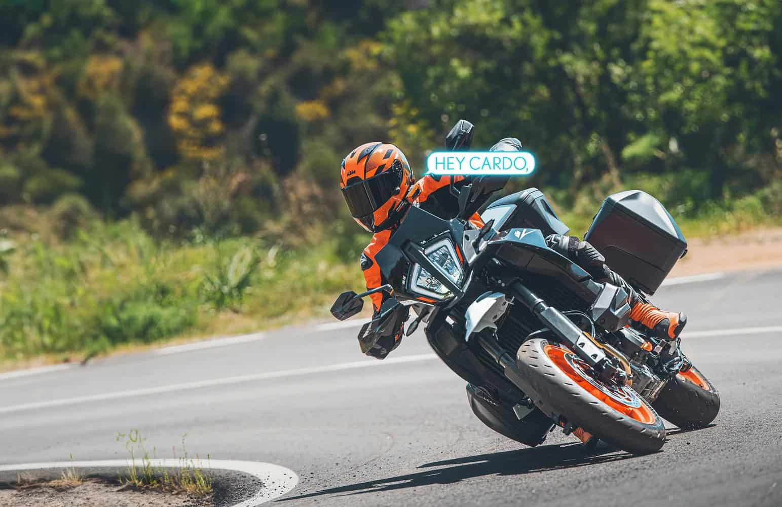 Motorcyclist leaning into a turn while using Cardo Packtalk Edge with voice command displayed as “Hey Cardo.”