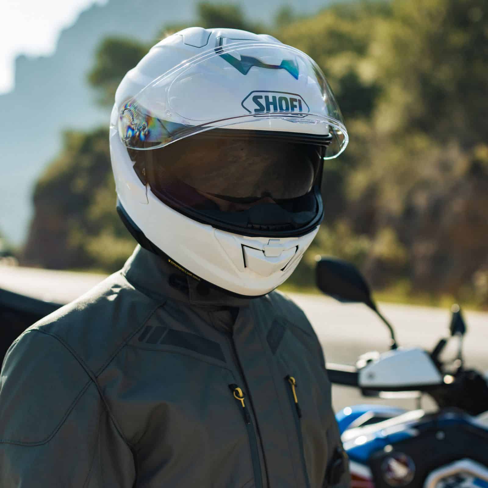 Rider wearing a white Shoei GT-Air 3 motorcycle helmet with drop-down sun visor, standing beside a touring bike.