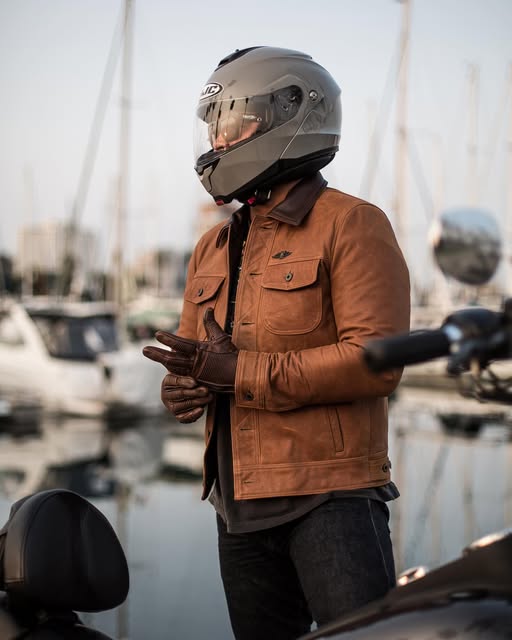 Rider wearing the HJC C91 modular helmet in matte silver, paired with a brown leather jacket, standing near a marina with boats in the background.