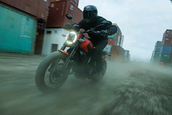 Rider wearing an HJC RPHA 12N helmet, cutting through dust on a motorcycle between shipping containers.