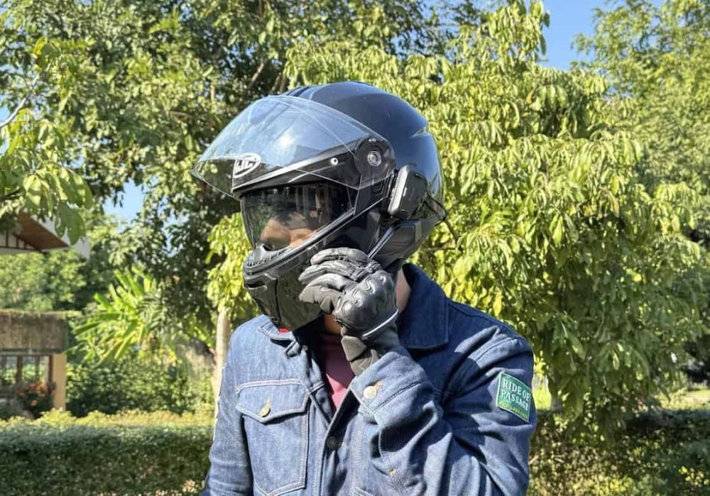 Rider adjusting a modular HJC i91 helmet with Bluetooth comms attached, standing outdoors in riding gloves and denim jacket.