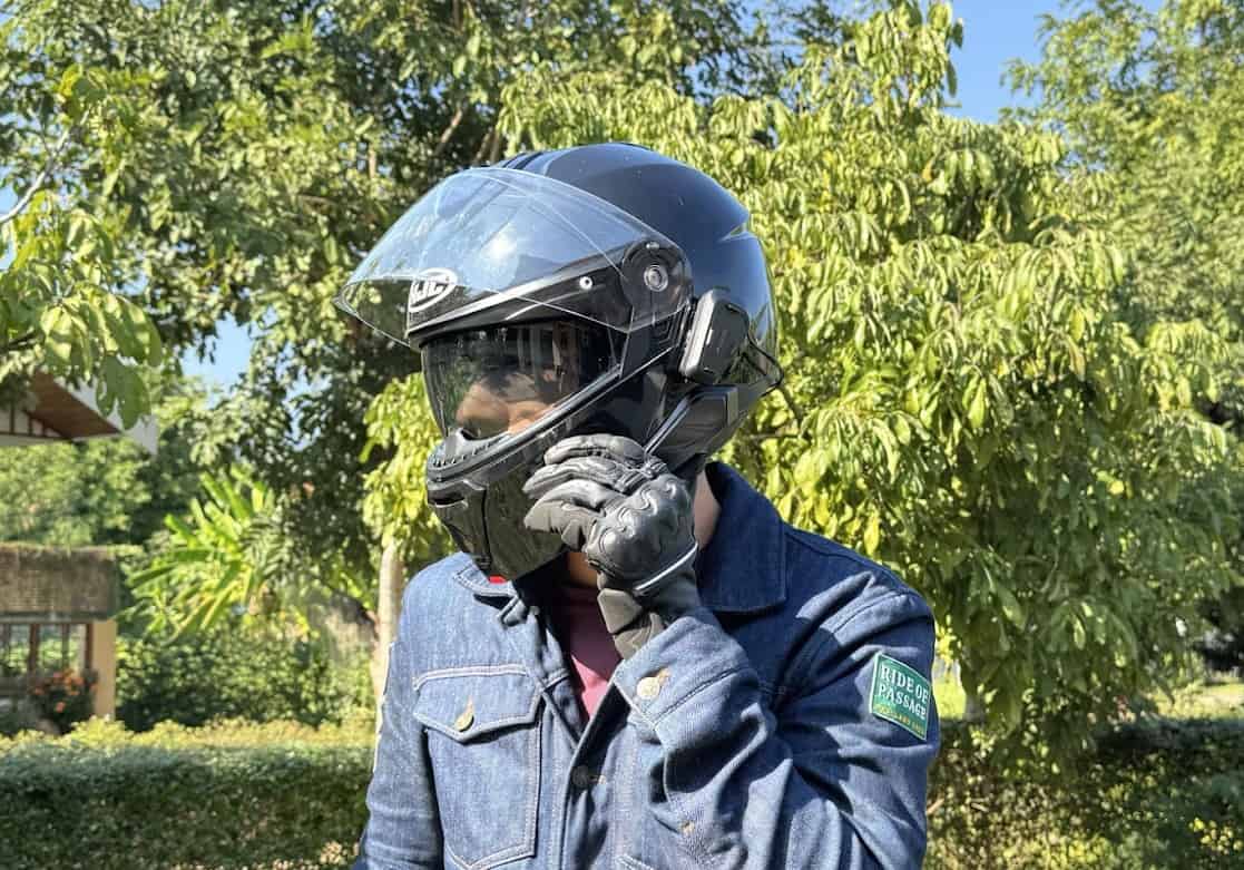 Rider adjusting a modular HJC i91 helmet with Bluetooth comms attached, standing outdoors in riding gloves and denim jacket.