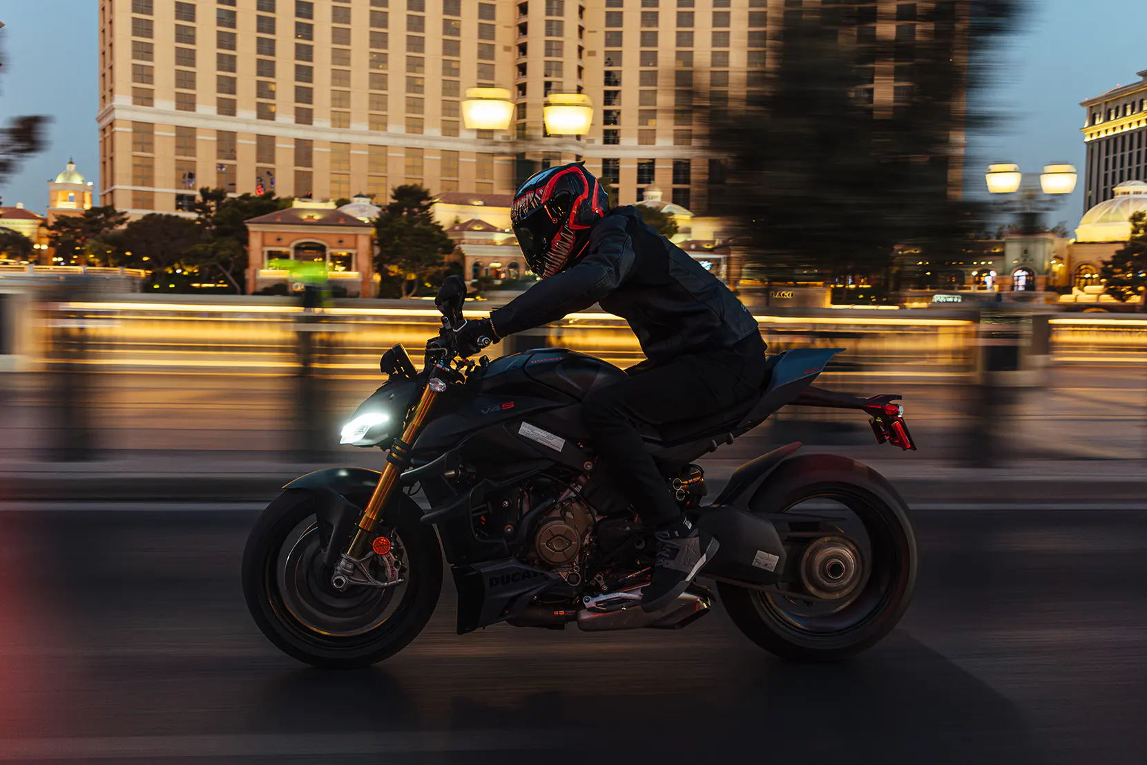 Motorcyclist riding a Ducati at night wearing a Venom-themed helmet with city lights blurred in the background.