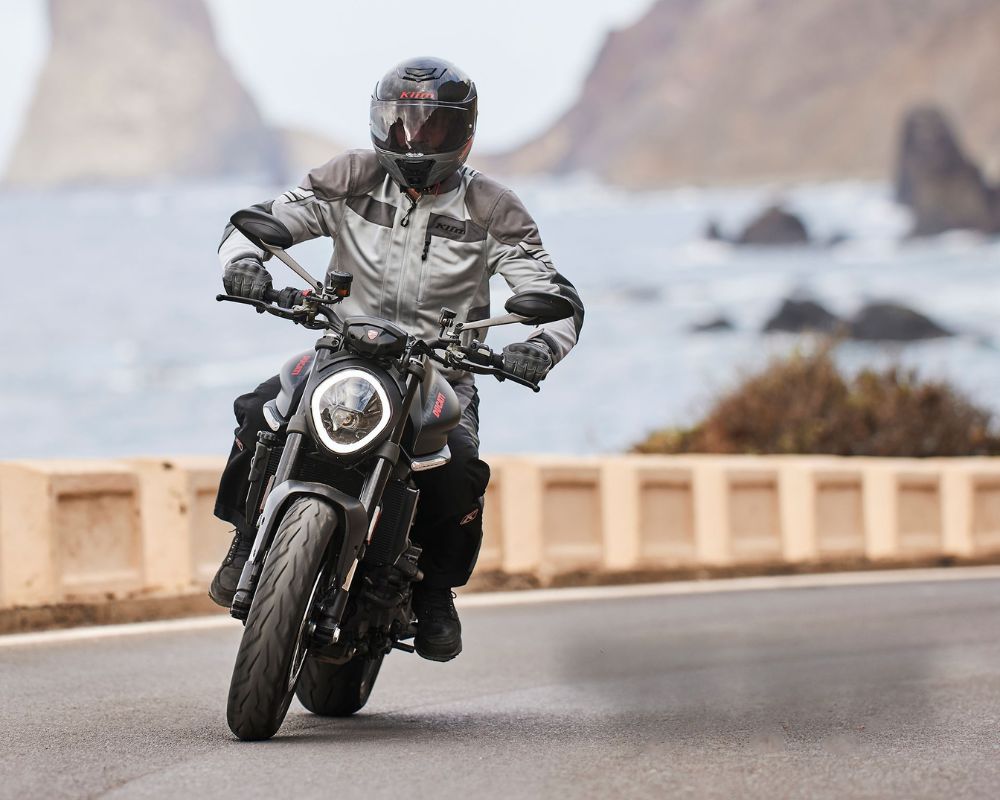 Rider wearing Klim Induction Pro jacket on a naked motorcycle along a coastal road