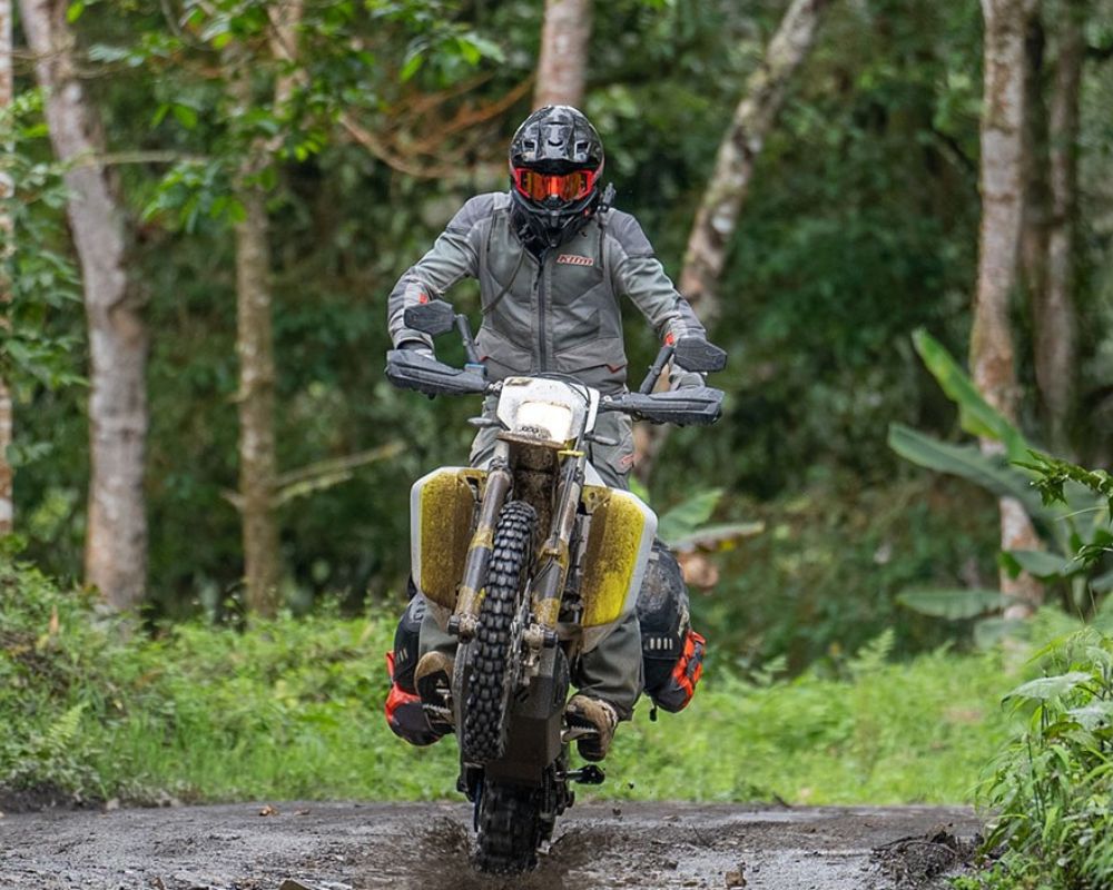 Rider wearing Klim Baja S4 jacket performing a wheelie on a dirt trail in tropical forest