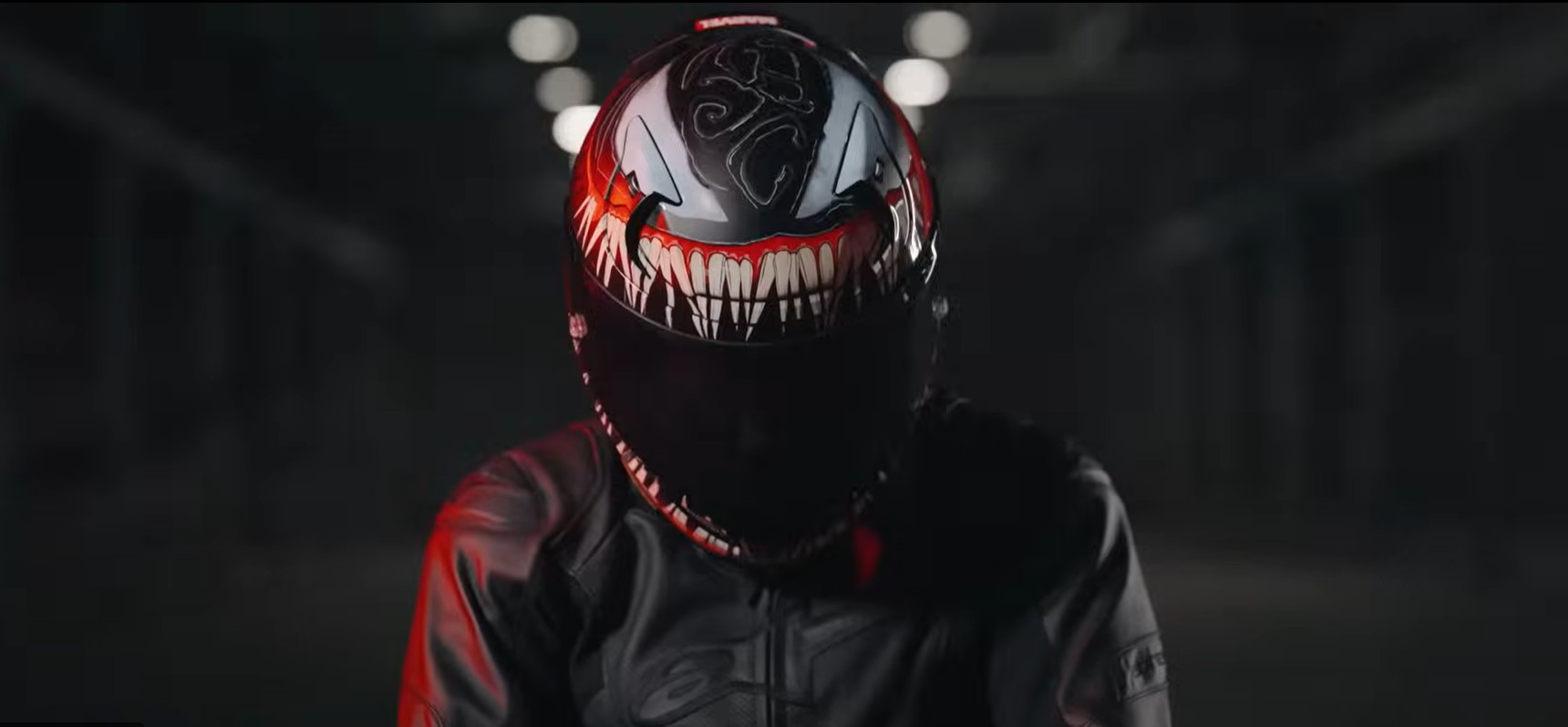 Rider in a dark setting wearing a Venom-themed motorcycle helmet and armored jacket with dramatic red lighting.