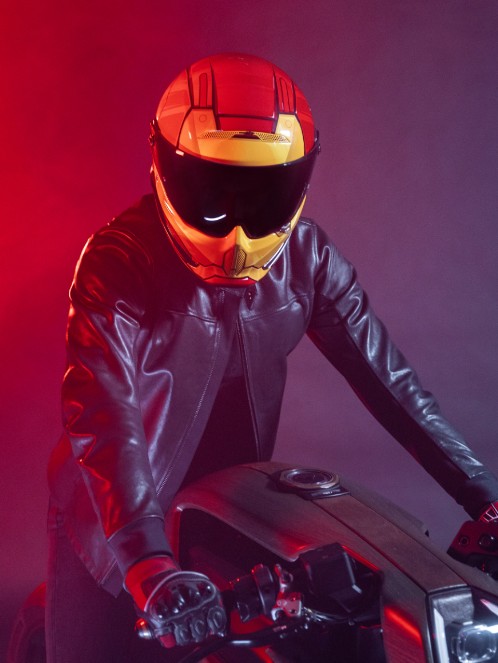 Rider wearing an Iron Man–themed motorcycle helmet with black visor, on a custom bike under red lighting.