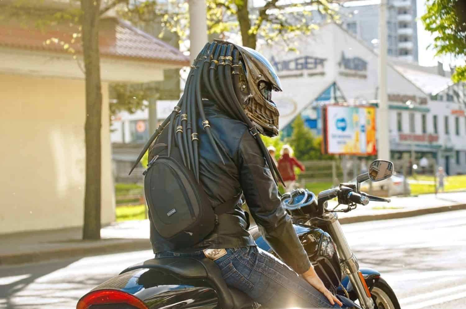 Motorcyclist wearing a Predator-style helmet with dreadlocks and sculpted faceplate, riding a cruiser through the city.