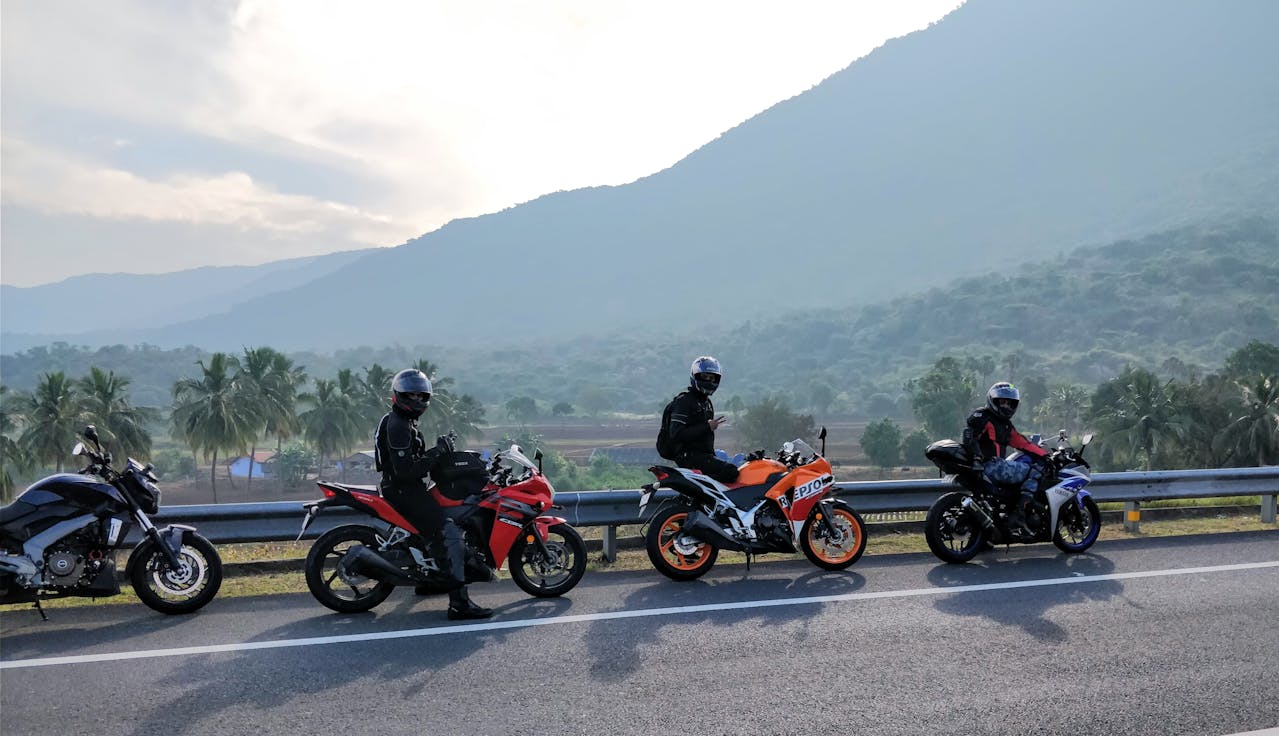 Group of motorcyclists stopped on the roadside with mountains in the background, wearing helmets and gear.