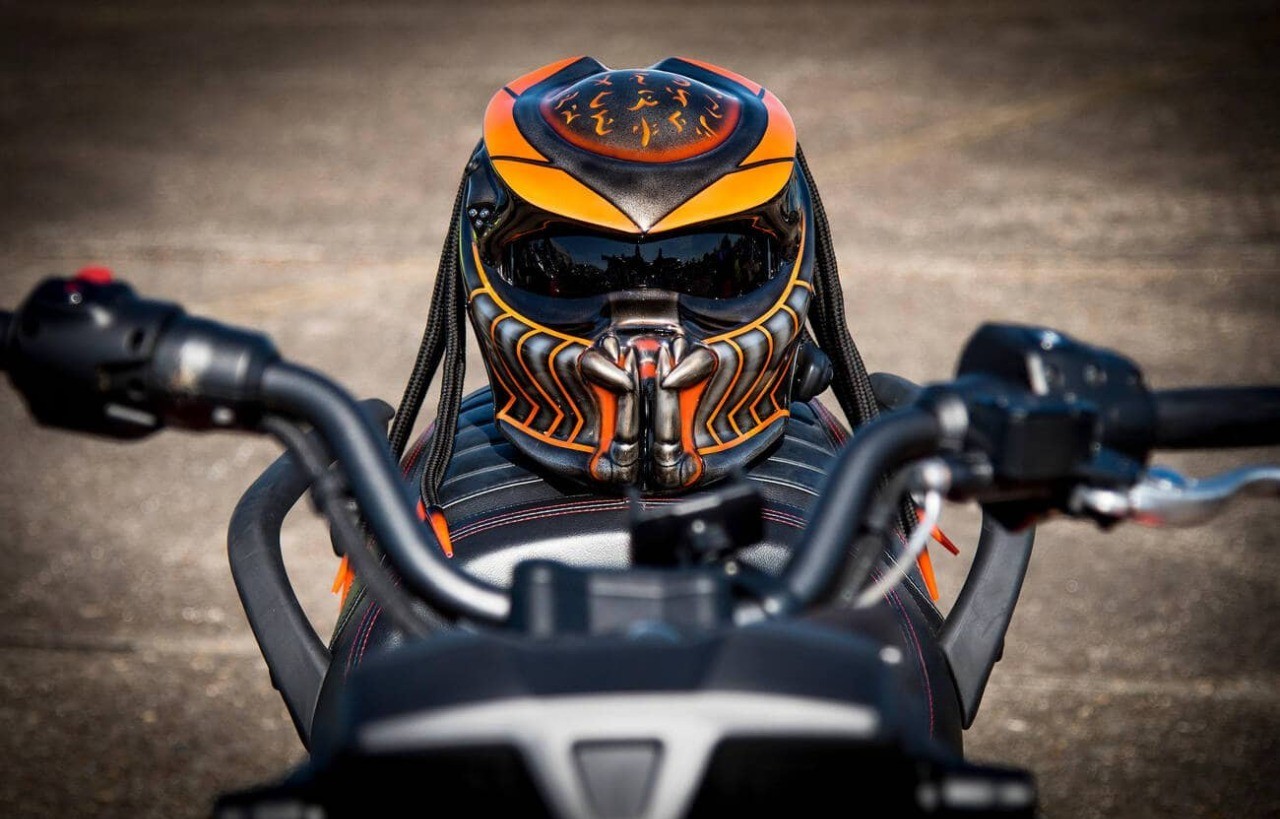 Predator-themed motorcycle helmet with orange and black airbrushed details and dreadlocks sitting on the seat of a parked motorbike.