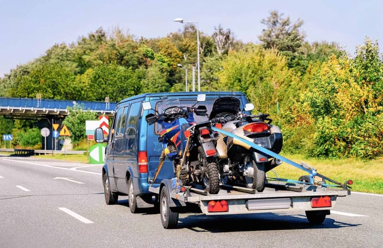 Blue van towing two motorcycles on an open double trailer along a road.