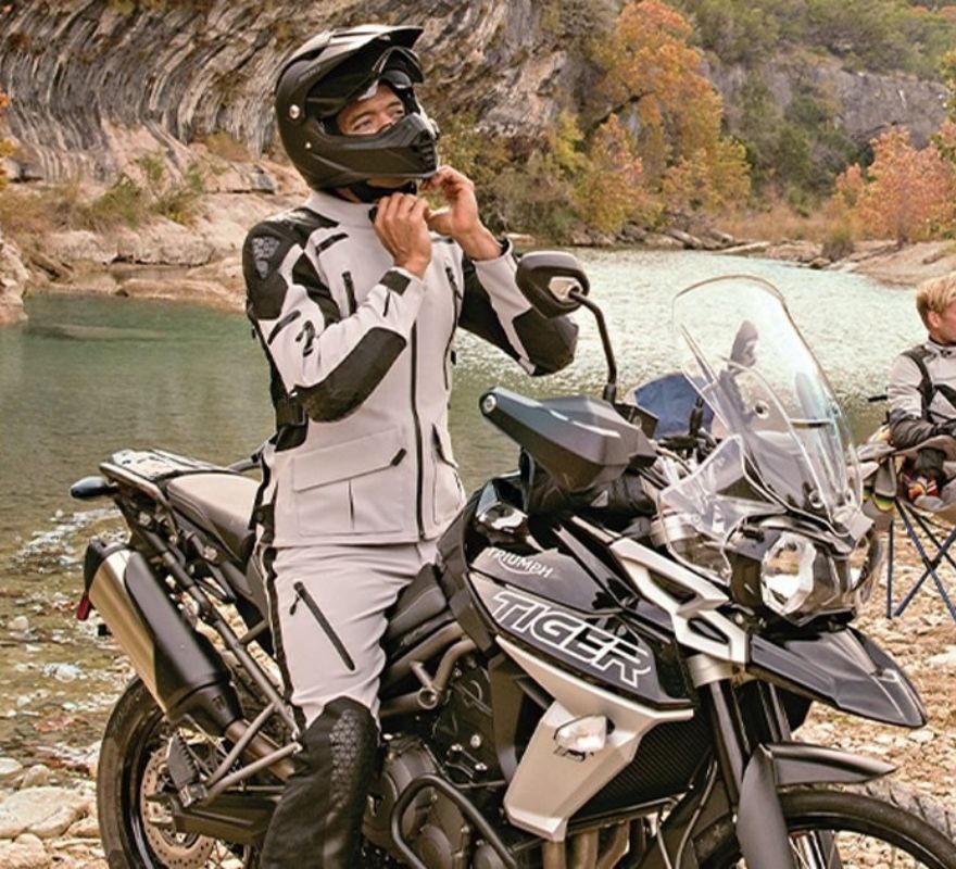 Rider in Sedici Garda jacket and pants on a Triumph Tiger by a river, adjusting helmet strap.