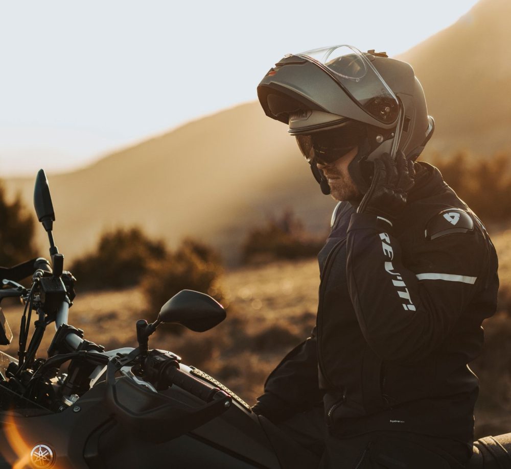 Rider wearing a Shoei Neotec 3 helmet with flip-up front open, sitting on a motorcycle at sunset