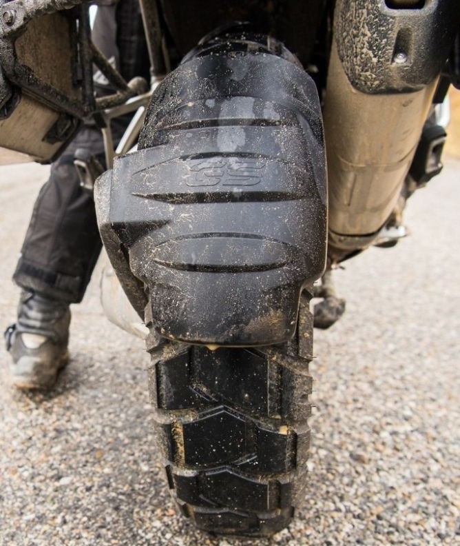 Close-up of a Shinko 705 dual-sport rear tire on a motorcycle, showing tread pattern with dirt and gravel