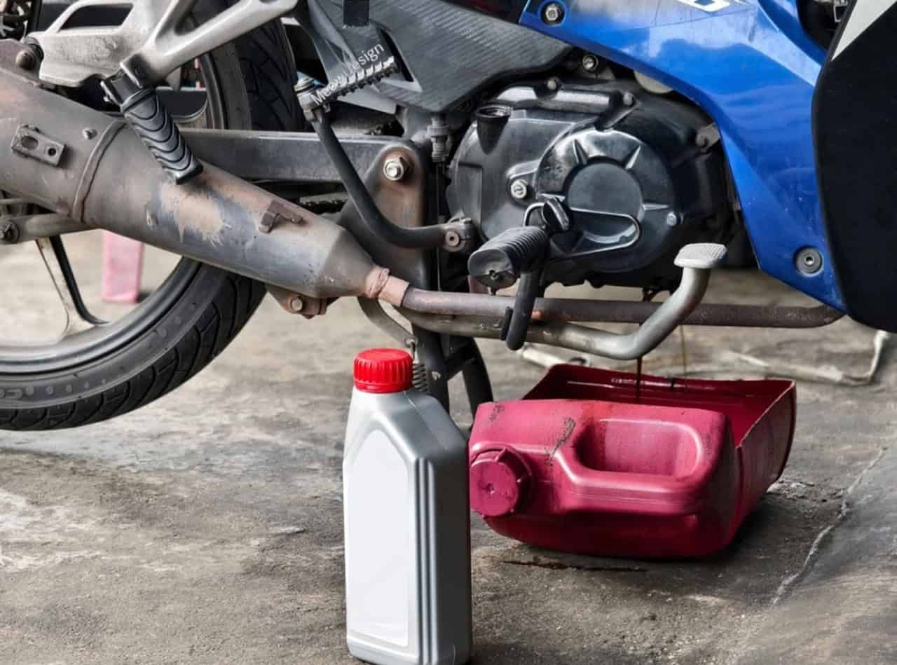 Motorcycle oil change in progress with used oil draining into a container and fresh oil bottle placed nearby