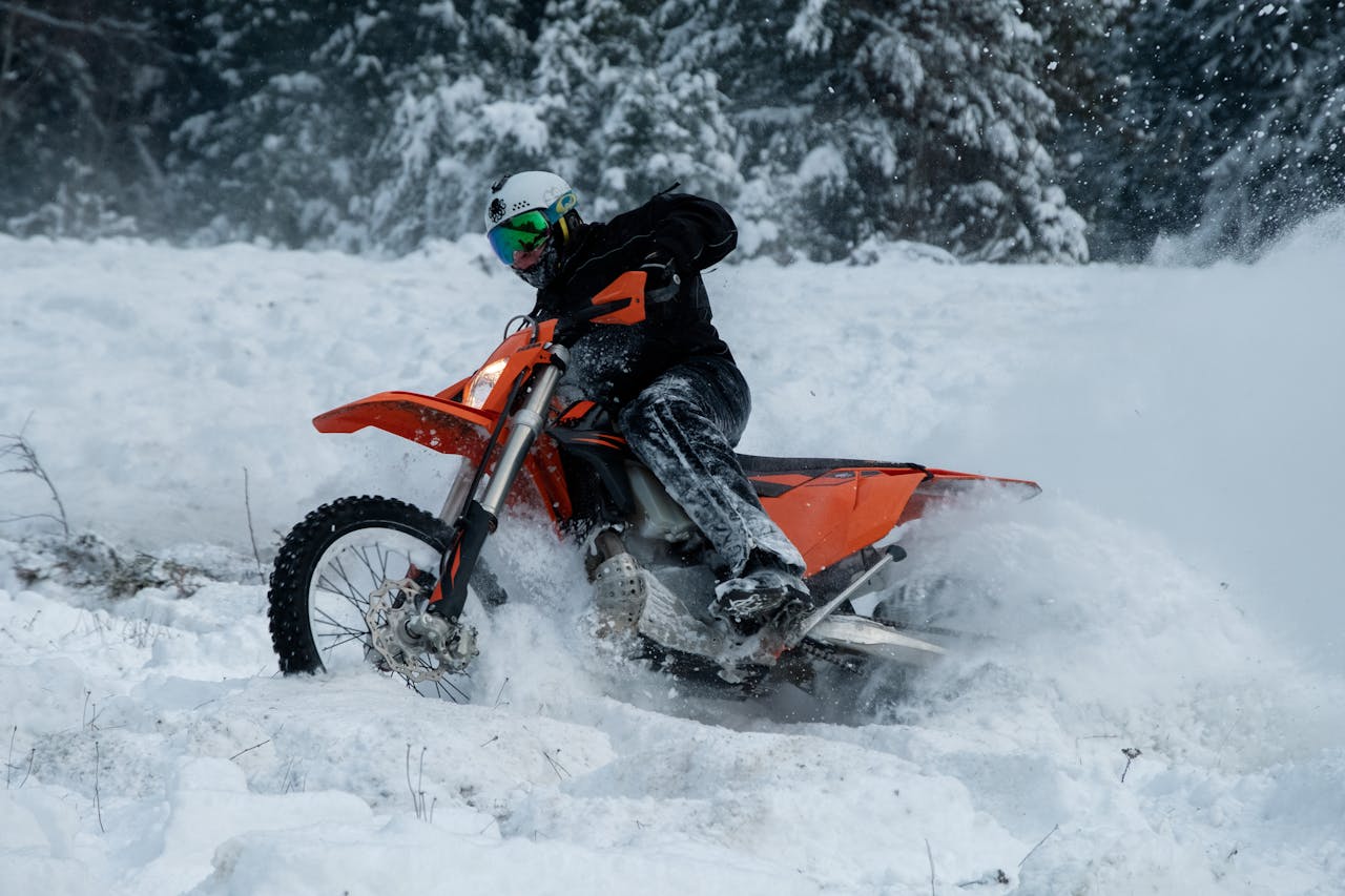Rider on orange dirt bike carving through snow in winter forest