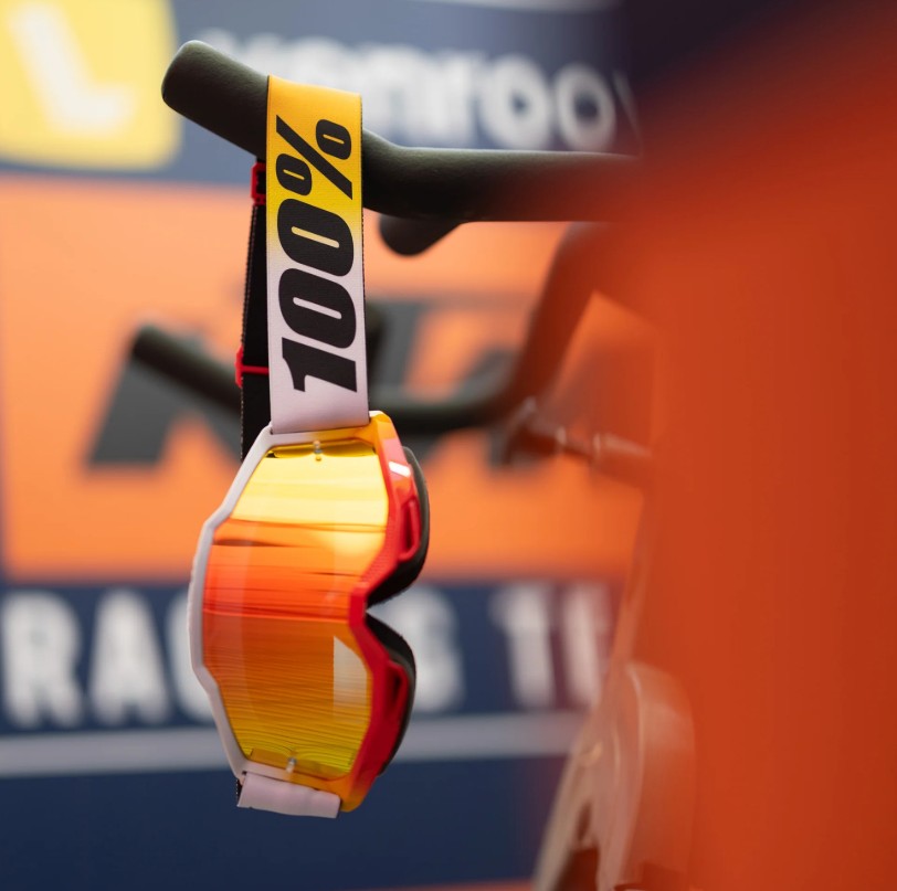 Close-up of orange-lensed motocross goggles with wide strap hanging from handlebars