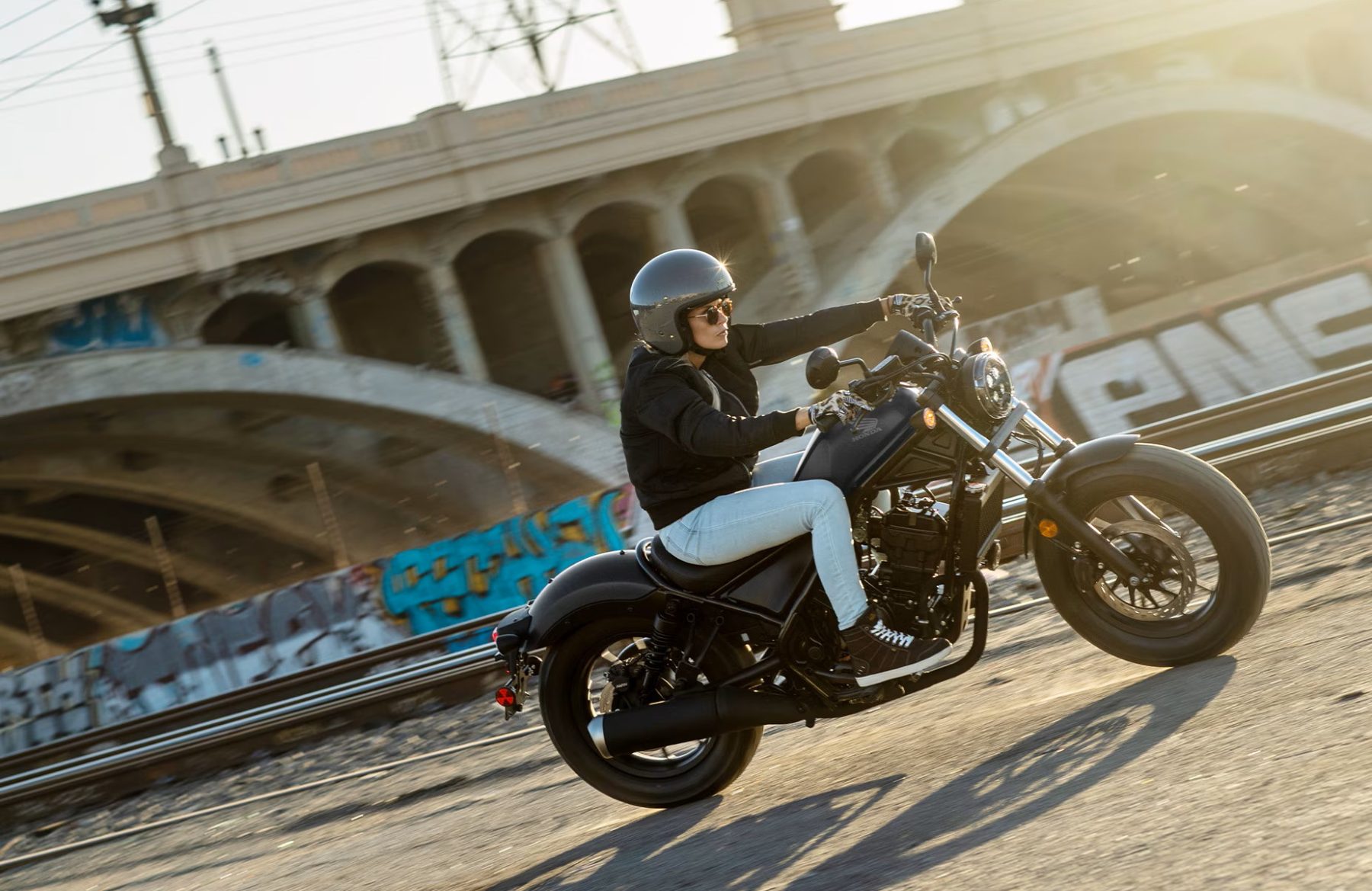 Woman riding a Honda Rebel 300 cruiser along an urban underpass at sunset