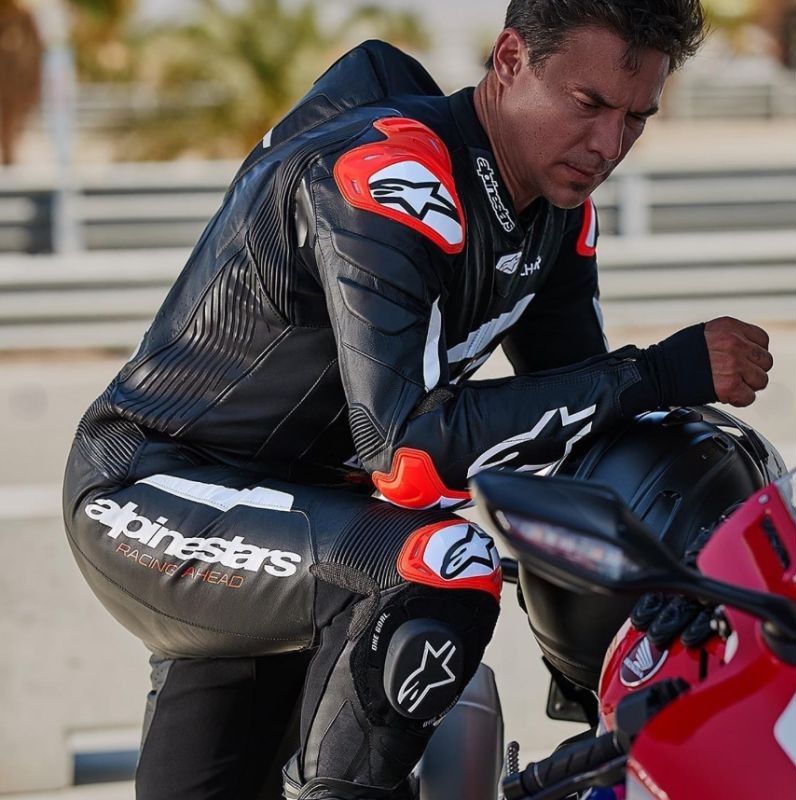 Close-up of a rider wearing an Alpinestars race suit, showing detailed cowhide leather panels, shoulder sliders, and reinforced stretch zones.
