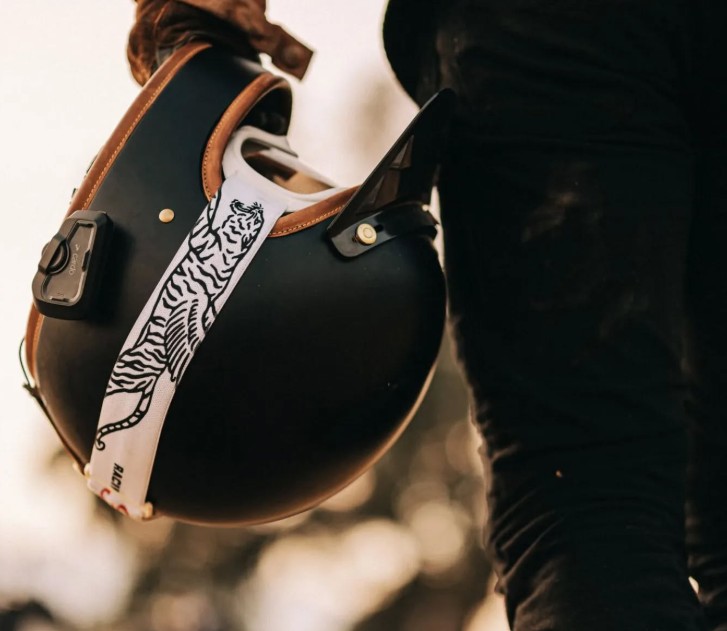 Close-up of a rider holding a matte black open-face helmet with a Cardo Freecom 4X communicator attached and a white goggle strap with a tiger print.