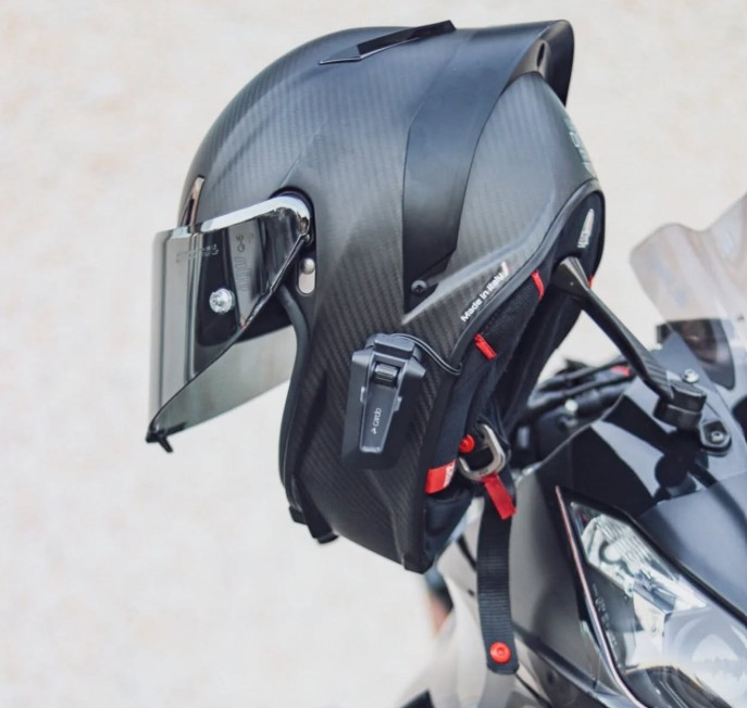 Close-up of a matte black carbon-fiber motorcycle helmet mounted on a bike, fitted with a Cardo Packtalk Neo communicator on the side.