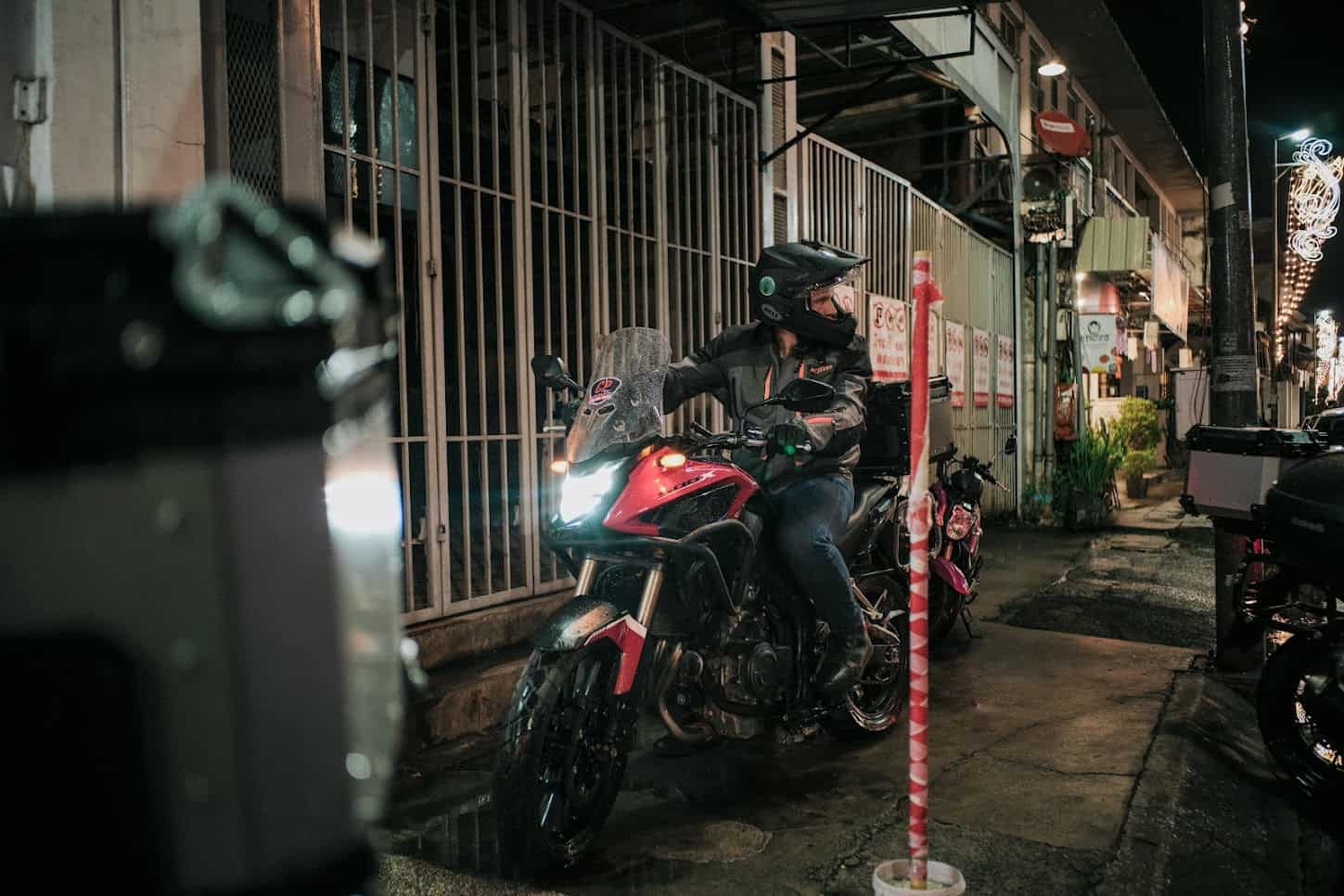 Motorcyclist wearing full riding gear and helmet, adjusting position while navigating a narrow city street at night