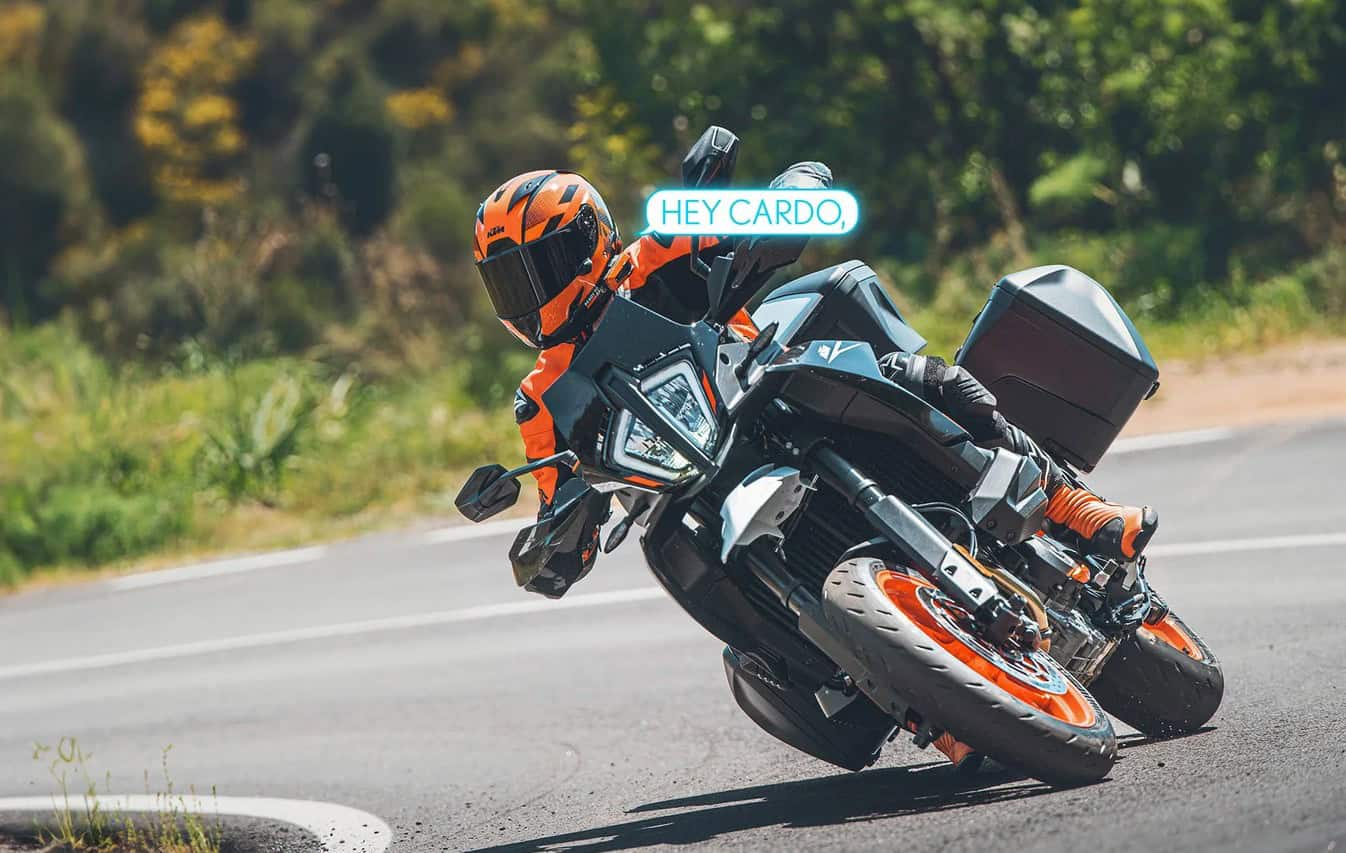 Motorcyclist leaning into a corner on a KTM wearing an orange helmet, using Cardo voice control with “Hey Cardo” command visible.