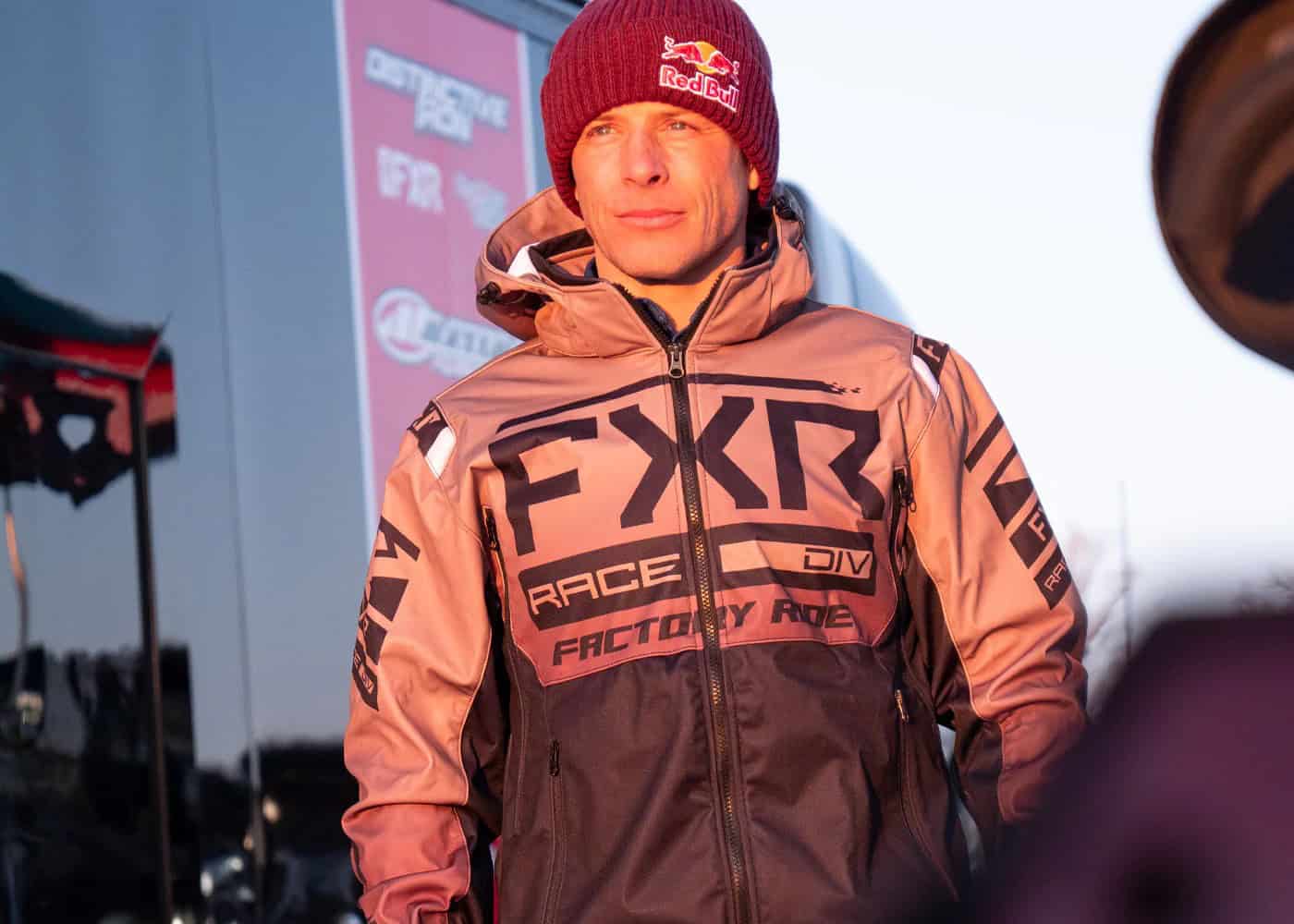 Rider wearing an FXR RRX Jacket in orange and black, standing outdoors in winter sunlight with Red Bull beanie and racing gear