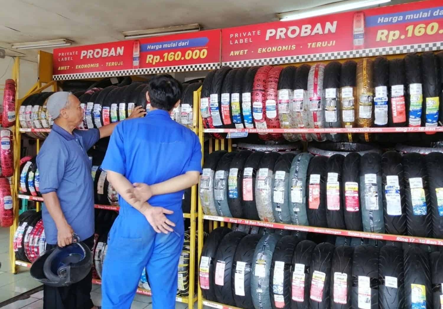 Two men inspecting motorcycle tires at a local shop with stacked shelves of various tire brands and sizes.