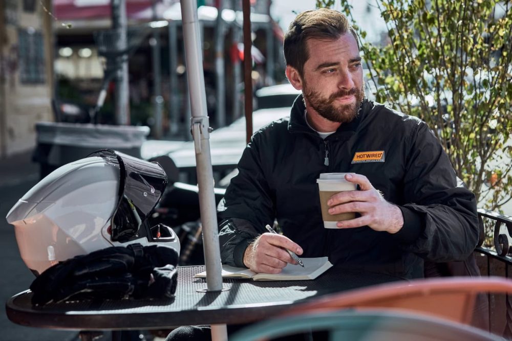 Rider wearing a Hotwired heated jacket, enjoying coffee outdoors, symbolizing built-in heating controls convenience