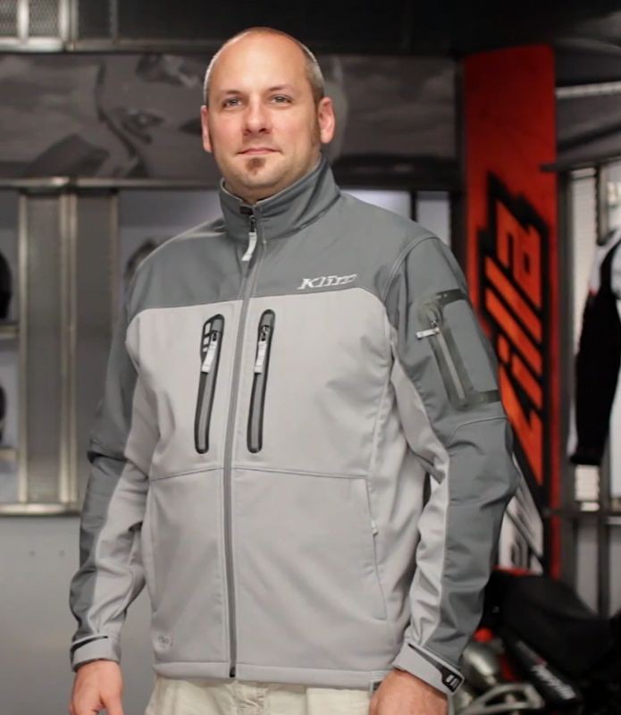 Man wearing Klim Inversion Jacket in a gear shop, showing the sleek gray soft-shell design with windproof zippers and high collar