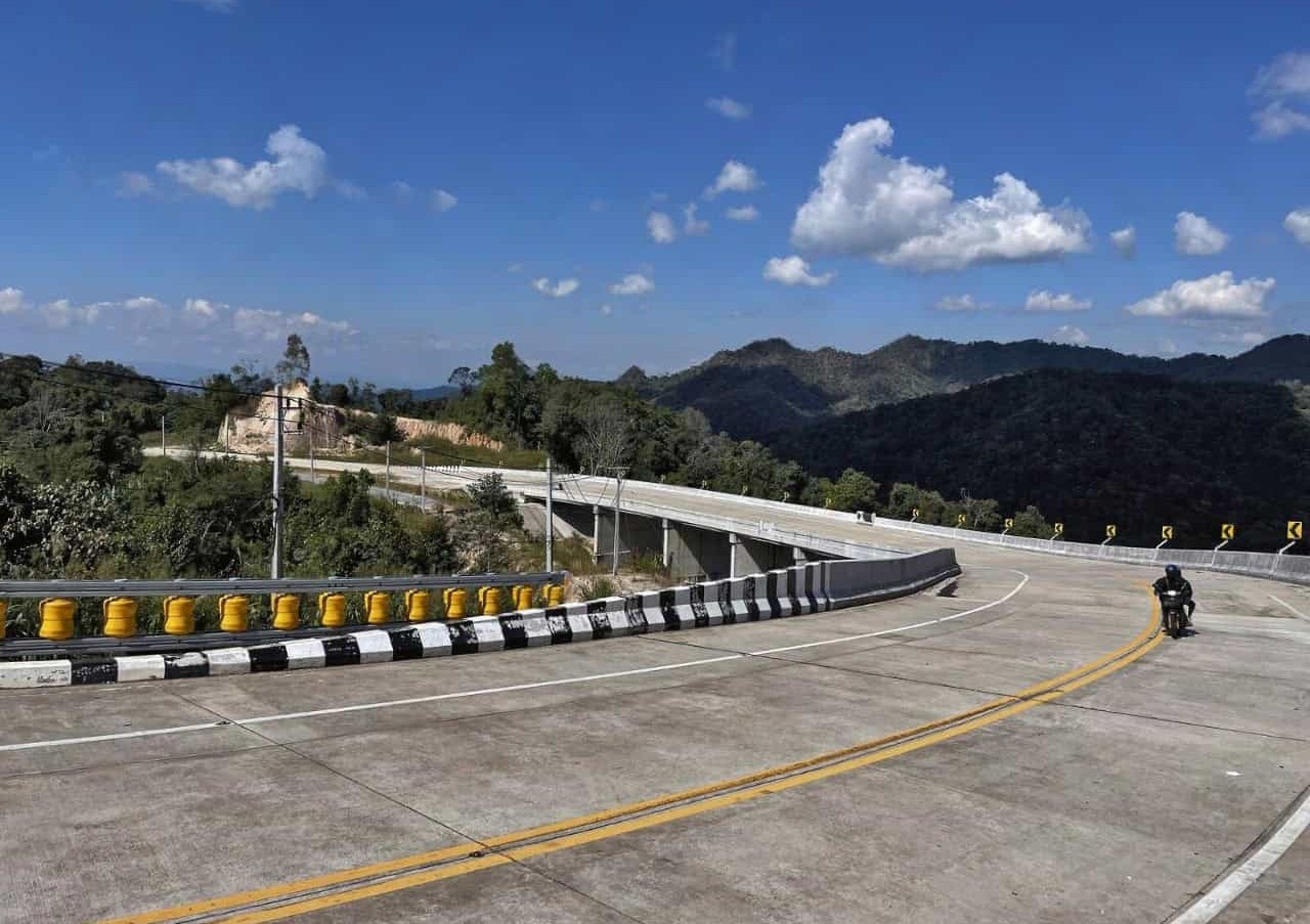 Motorcyclist riding along the winding Mae Hong Son loop highway in northern Thailand, surrounded by lush mountains and clear blue skies