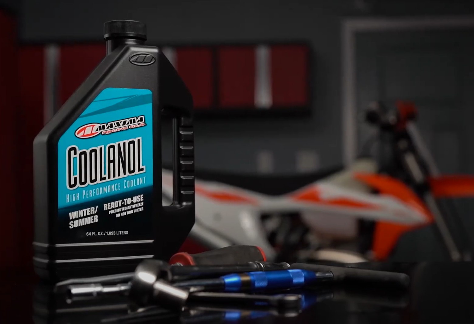 Maxima Coolanol High Performance Coolant bottle on a workbench beside tools and a dirt bike in a garage