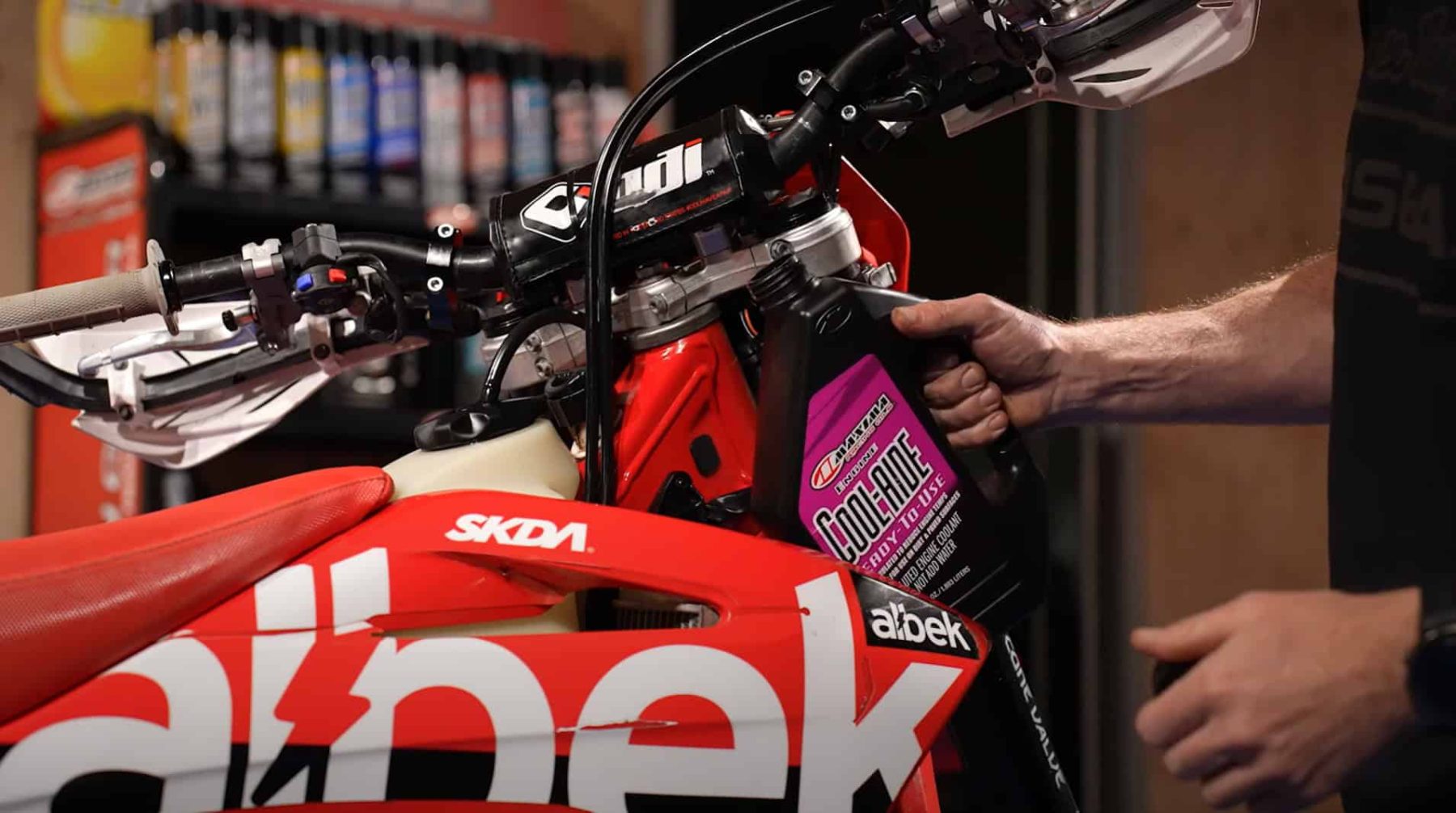 Mechanic pouring Maxima Cool-Aide Coolant into a red dirt bike during maintenance in a garage