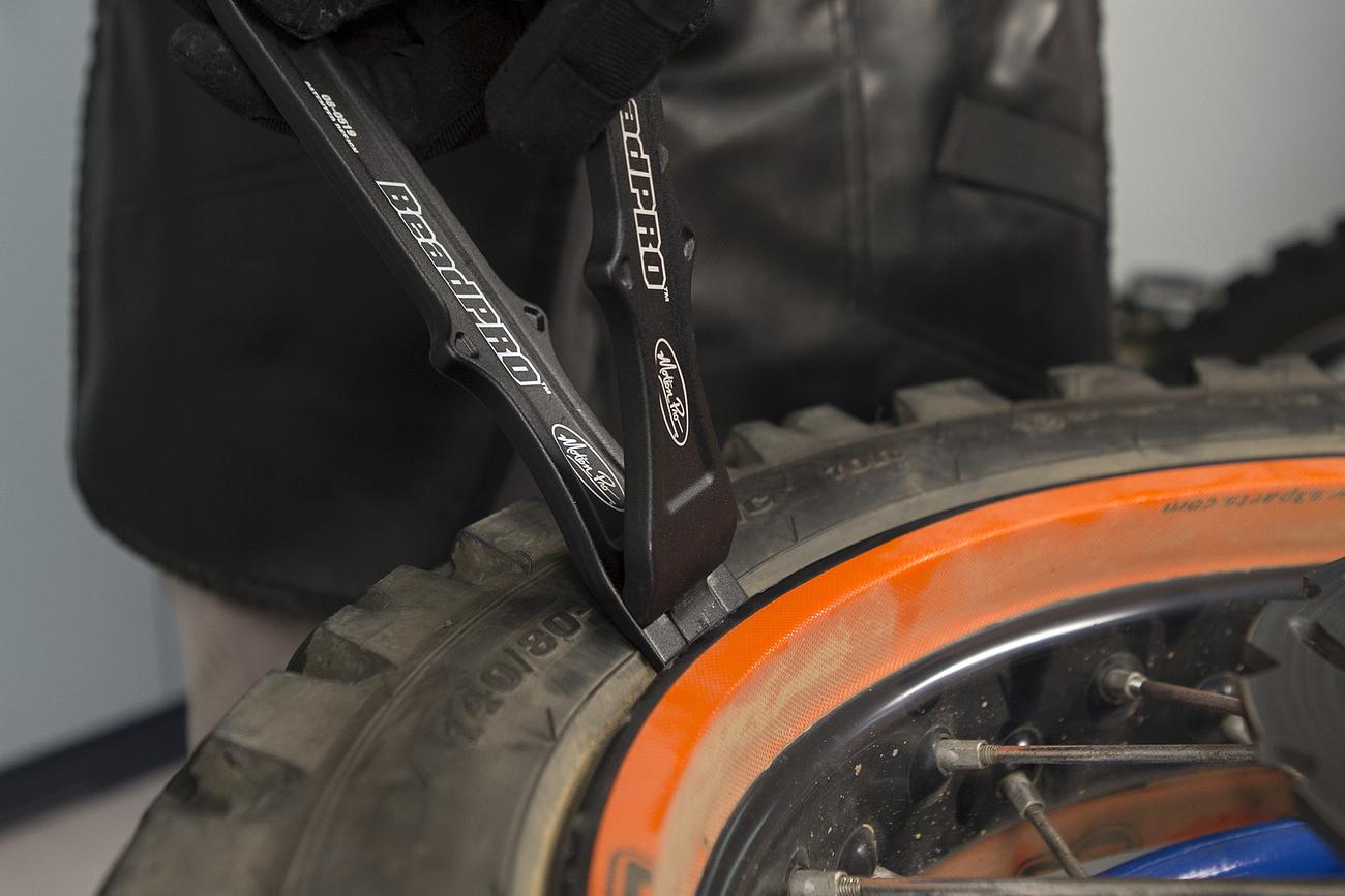 Close-up of Motion Pro BeadPro levers breaking a motorcycle tire bead on an orange-rimmed wheel.