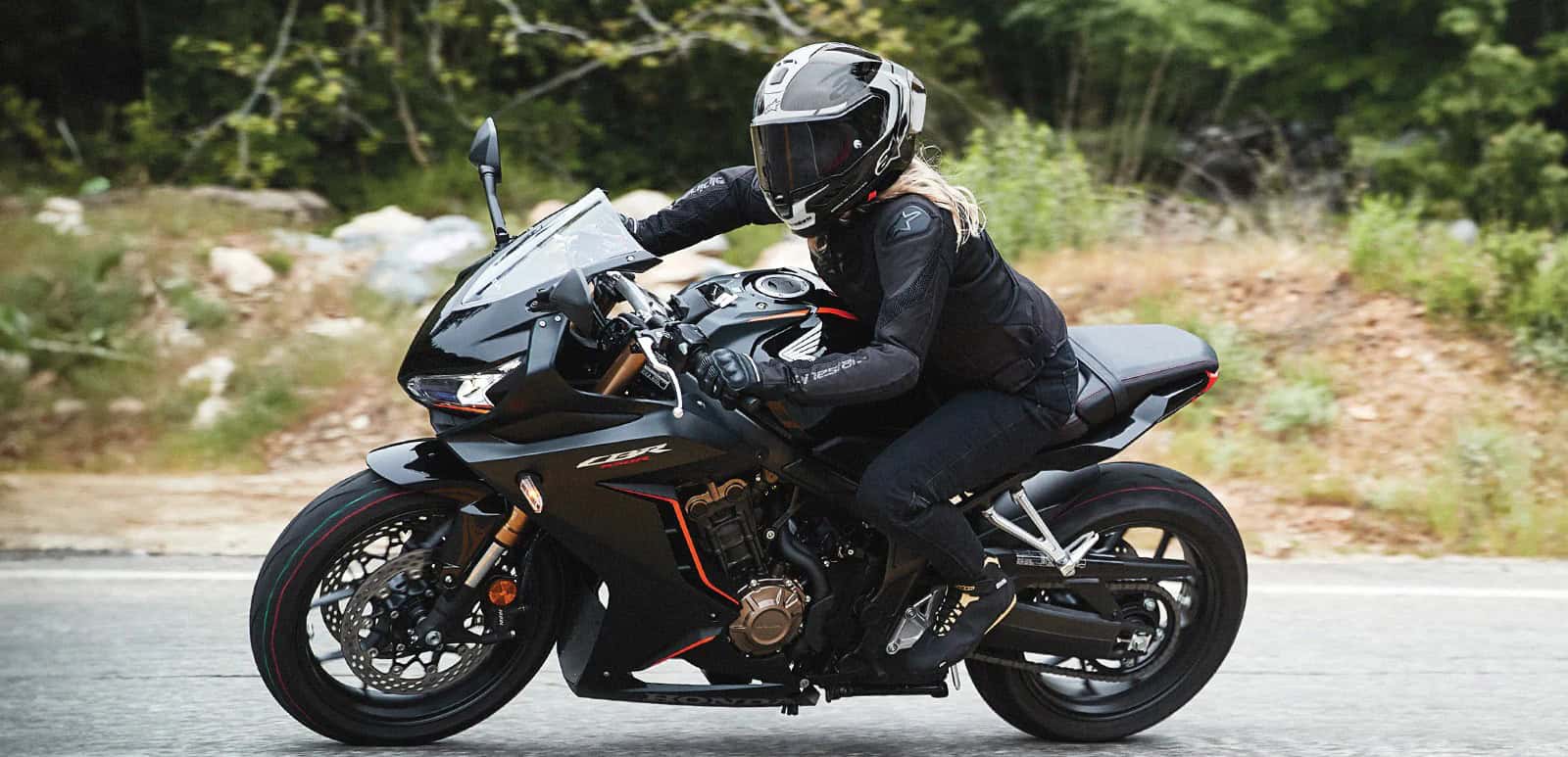 Woman riding a black sportbike wearing full black gear and helmet, showcasing the Alpinestars Stella T-GP Plus R V4 Airflow Jacket on a twisty road.