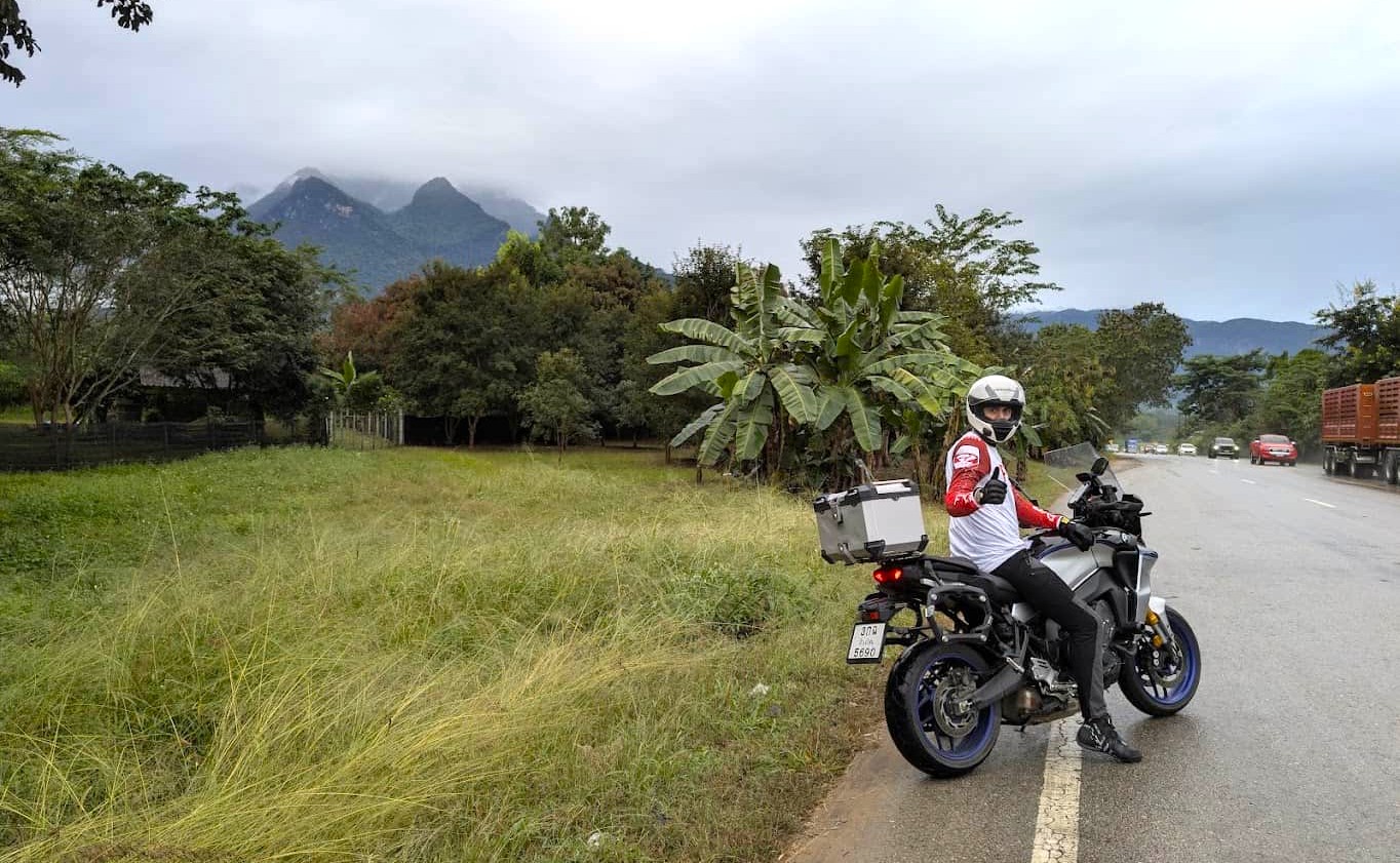 Motorcyclist wearing a full-face helmet and protective gear on a touring bike in rural Thailand, giving a thumbs-up beside the road