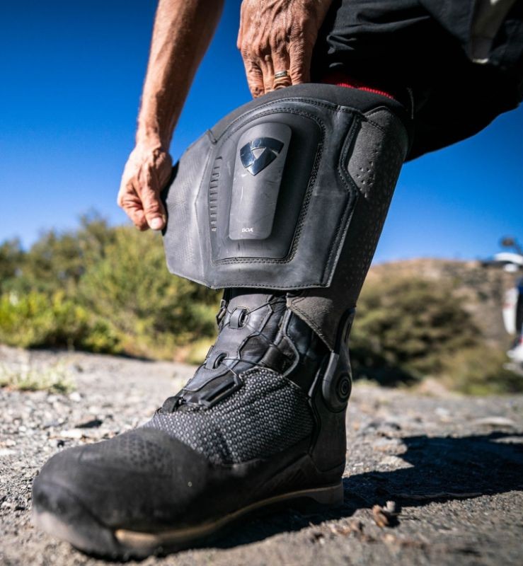 Rider tightening the BOA dial on a REV’IT! Expedition GTX boot, showing advanced closure and shin protection detail.